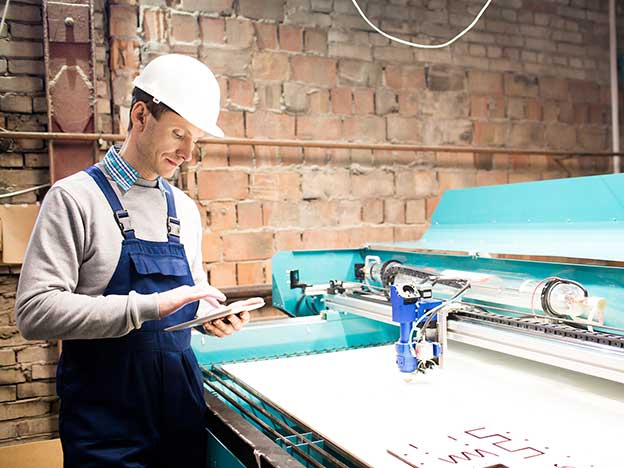 A manufacturing engineer wearing a hardhat and factory apron works with a tablet to operate an insulation cutting table.